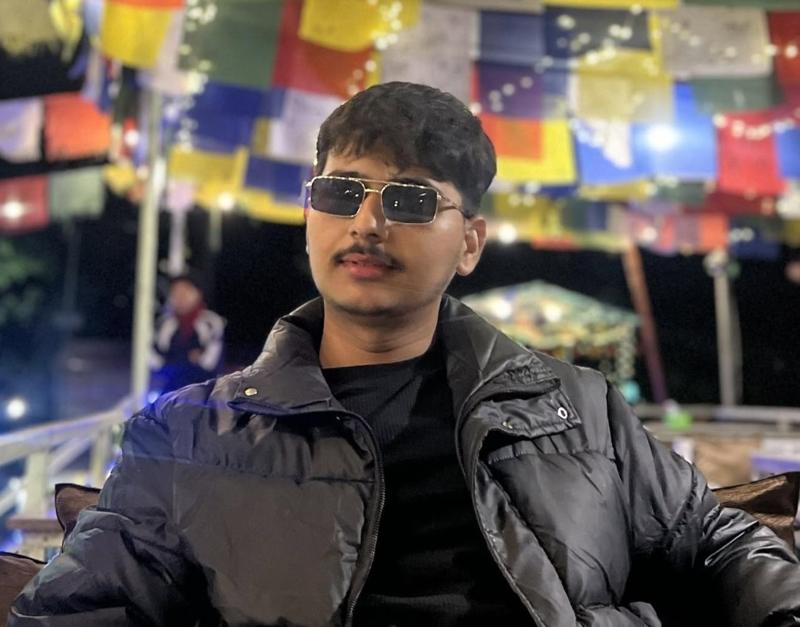 Arjun Rizal wearing sunglasses and a black jacket in front of colourful prayer flags at night